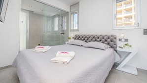 A modern bedroom with a large bed, a glass shower enclosure, and a view of the building's balconies.