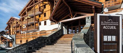 Una cabaña de estilo chalet con una escalera de piedra que conduce a una puerta de madera con la leyenda 'ACCUEIL'.