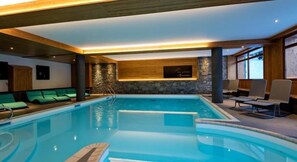 Piscina interna com parede de pedra, vigas de madeira e espreguiçadeiras.