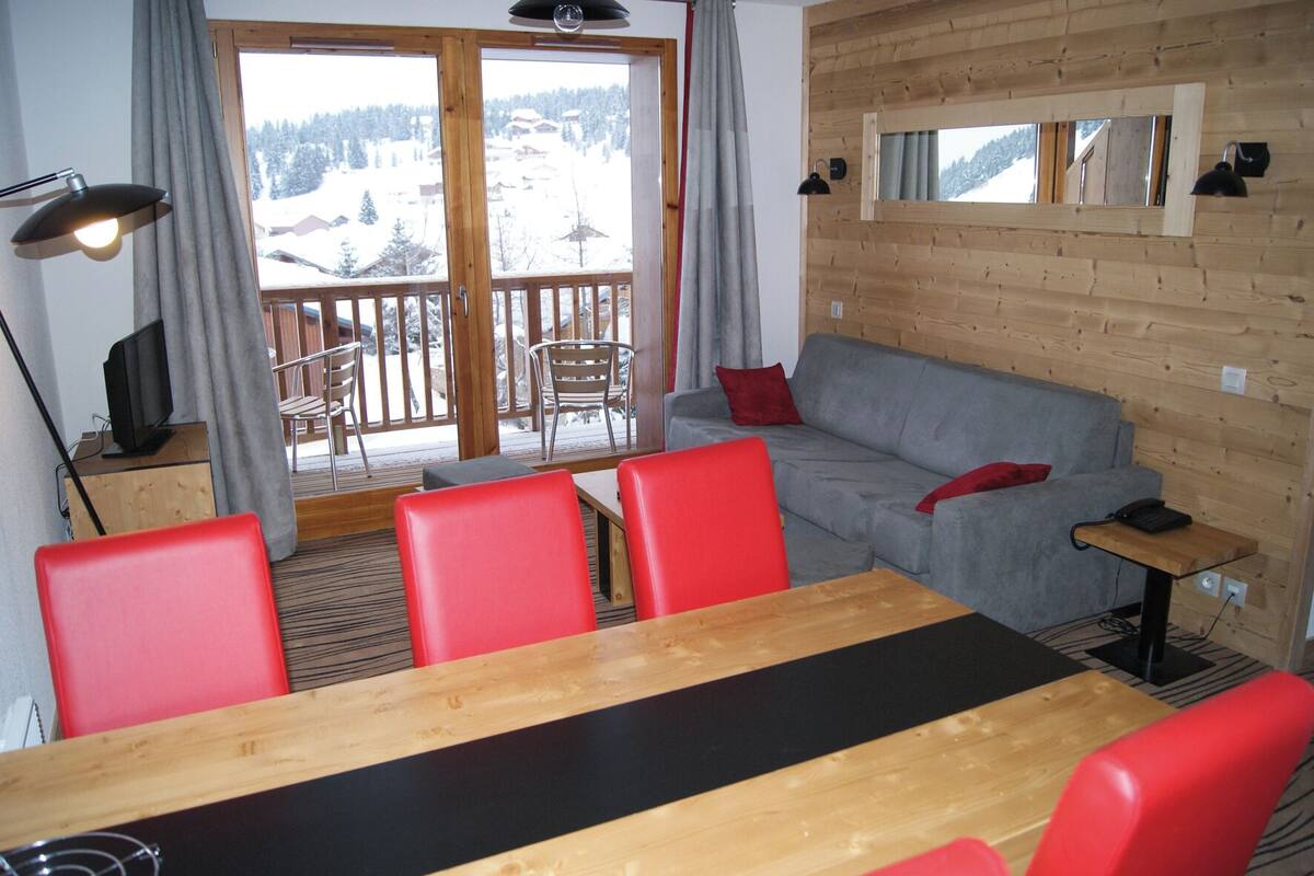 A room with a wooden interior, a grey sofa, red chairs, a wooden table, a TV on a wooden cabinet, and a balcony with a view of a snowy landscape.