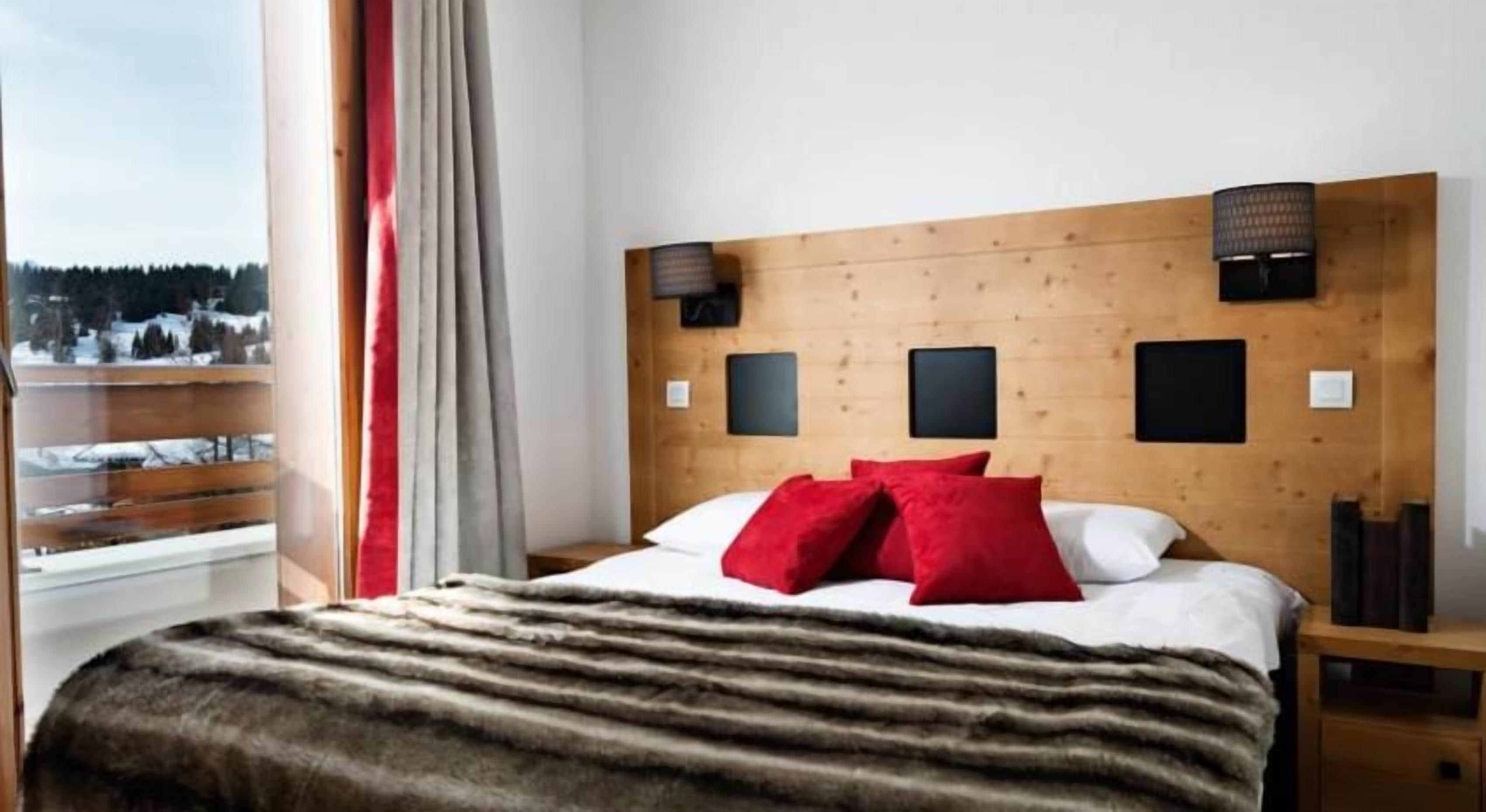 Habitación de hotel con cabecera de madera, una cama grande con almohadas rojas y blancas, un plaid de piel y vistas a un paisaje nevado a través de una ventana.