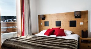 Habitación de hotel con cabecera de madera, una cama grande con almohadas rojas y blancas, un plaid de piel y vistas a un paisaje nevado a través de una ventana.