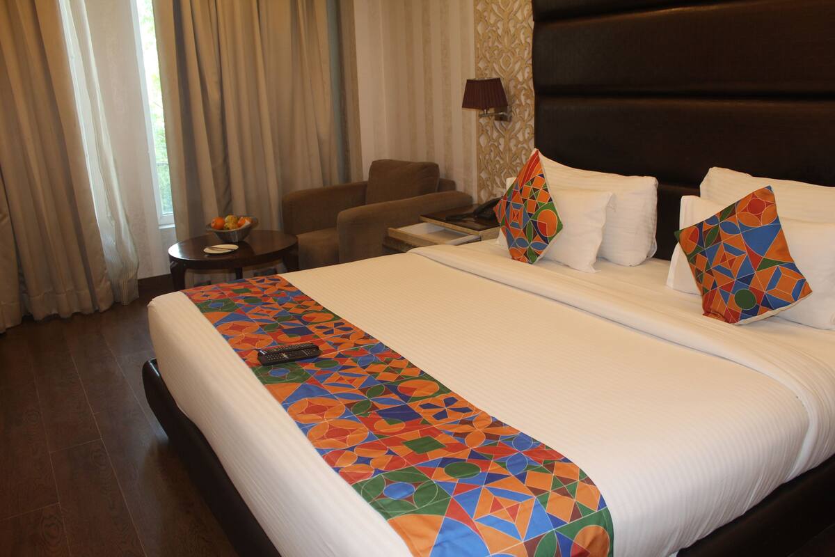 A hotel room with a large bed, a chair, a small table with a bowl of fruit, and a lamp on the wall.