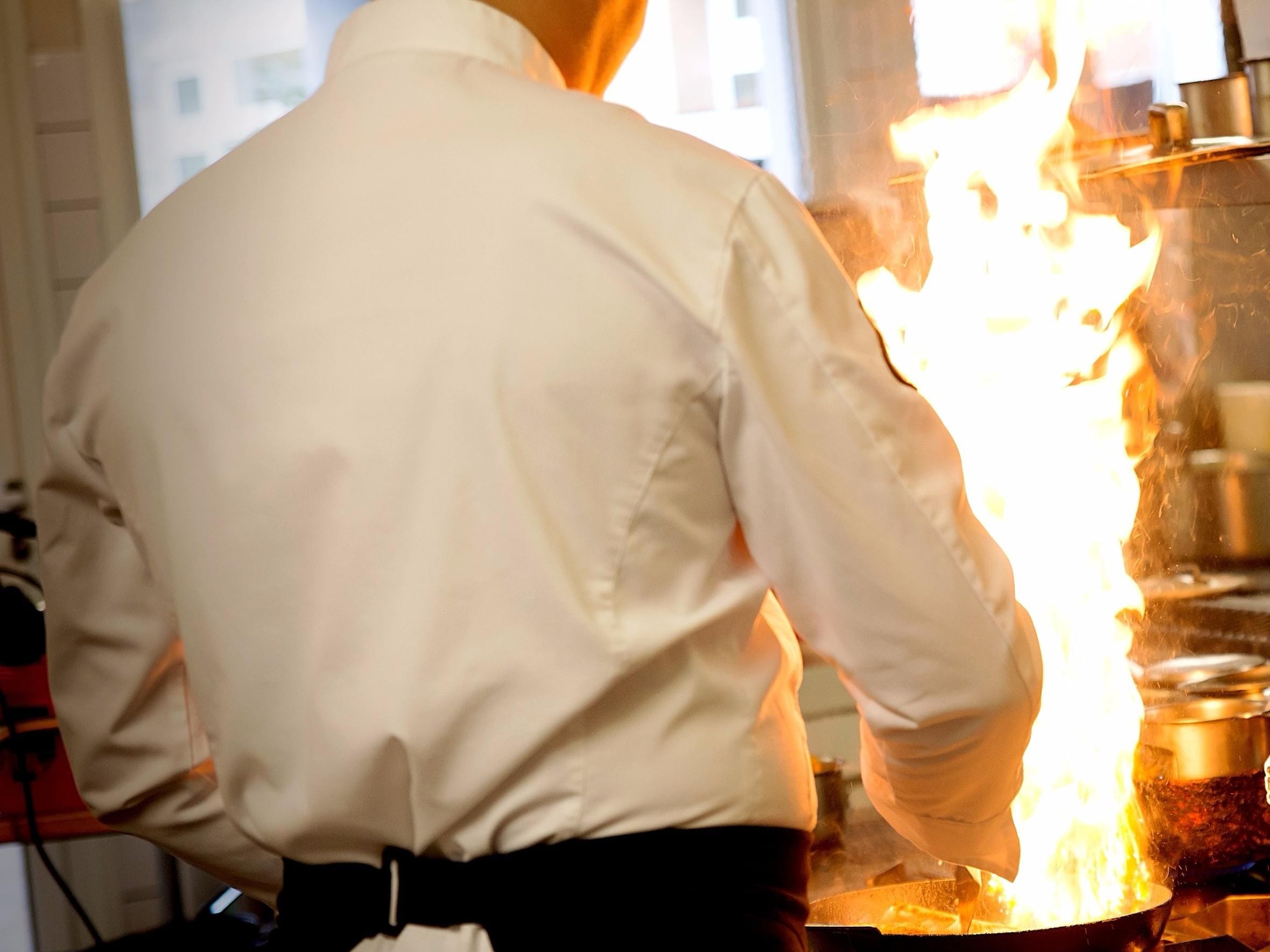 Eine Person in weißem Hemd und schwarzer Hose steht vor einem Grill mit Flammen.