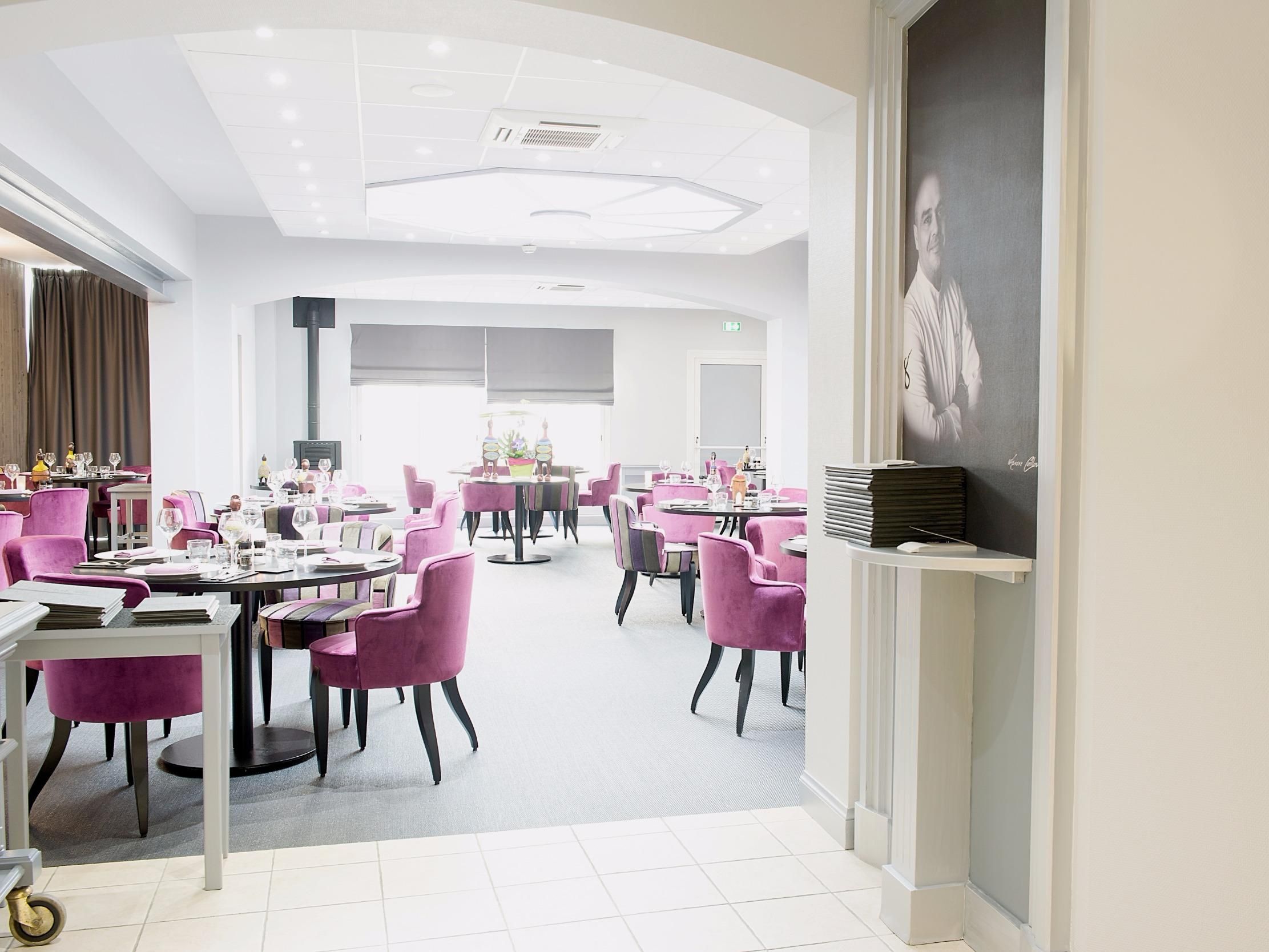Un espace de restauration moderne avec des tables et des chaises rondes, un plafond doté d’un éclairage encastré et un grand portrait accroché au mur.