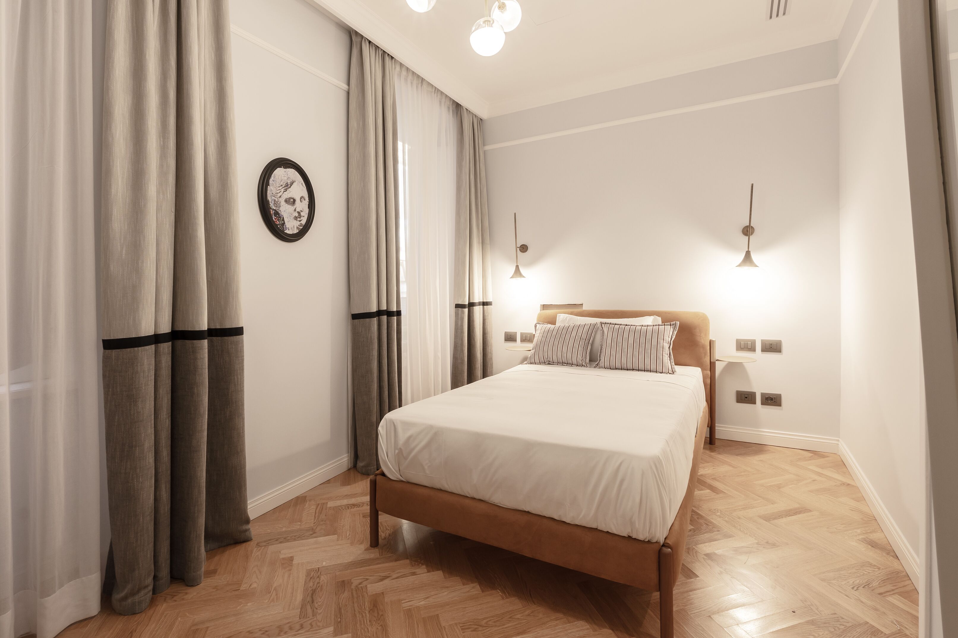 A bedroom with a wooden bed, white bedding, a headboard, two bedside lamps, and a large window with sheer curtains.