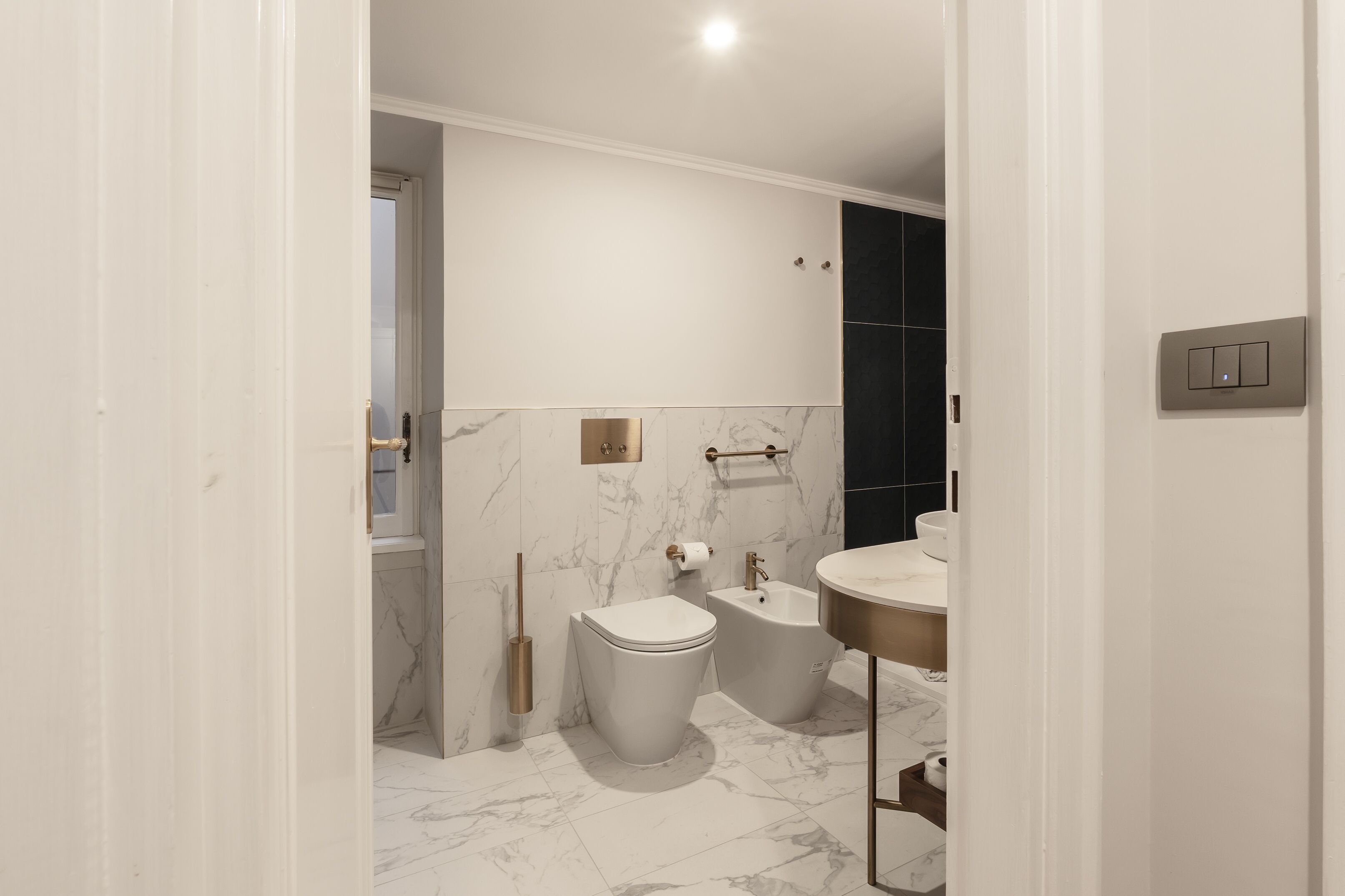 Un baño moderno con inodoro, bidé y lavamanos, suelo de mármol y pared blanca con interruptor de luz.