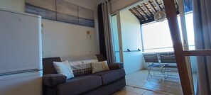 A living room with a sofa, a table, and a chair. There is a window with curtains, a ceiling fan, and a wall-mounted light fixture. - Appartement Tout Confort Dans Résidence Pierre & Vacances (Grimaud)