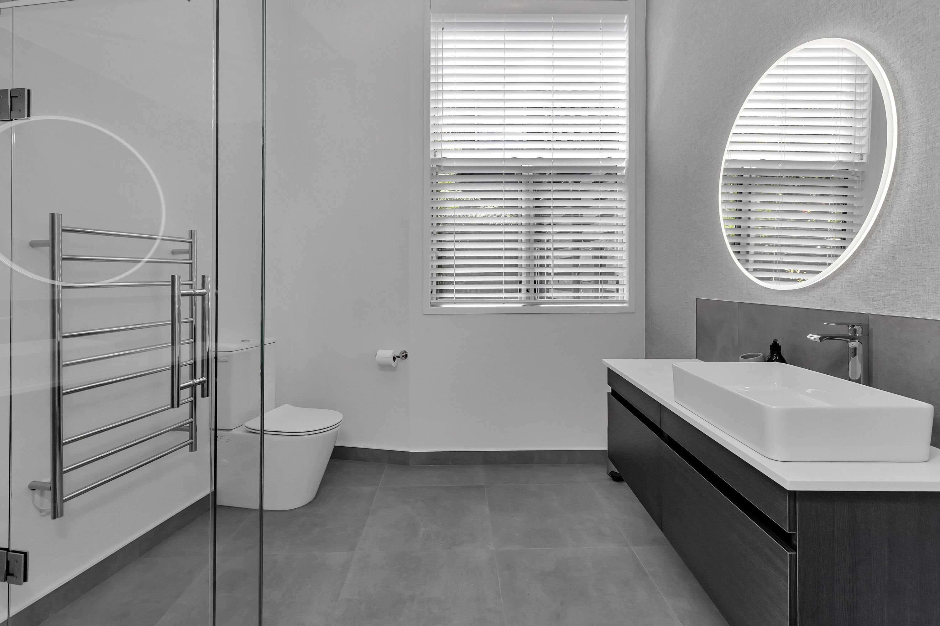 A modern bathroom with a glass shower enclosure, a white toilet, a vanity with a large sink, and a window with blinds.