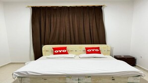 A single bed with a white mattress and two red pillows with "OYO" branding, a brown curtain, and a small wooden nightstand. - Super OYO 144 Al Tawasi Furnished Apartments (Salalah)