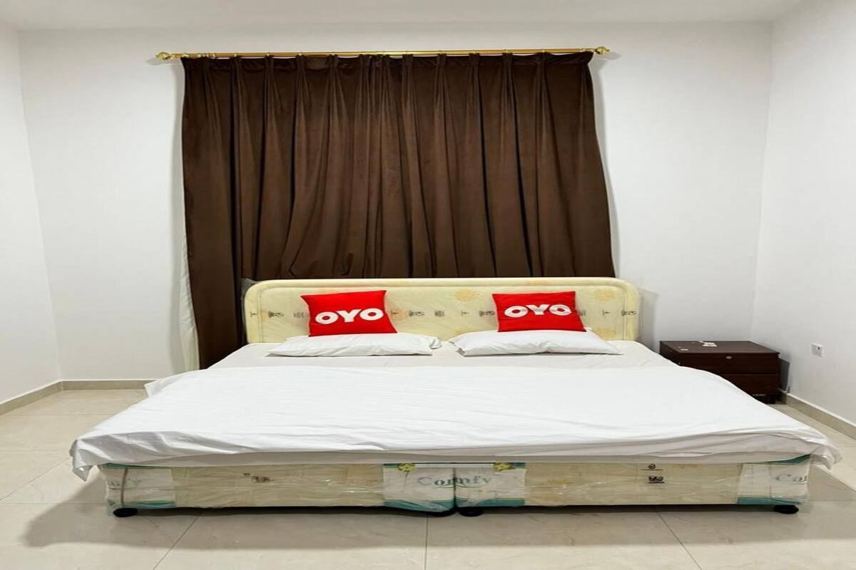 A single bed with a white mattress and two red pillows with "OYO" branding, a brown curtain, and a small wooden nightstand.