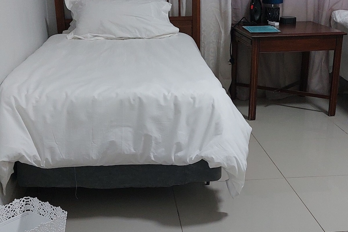 Una cama bien hecha con ropa blanca, un cabecero de madera y un cubo de basura blanco a su lado.