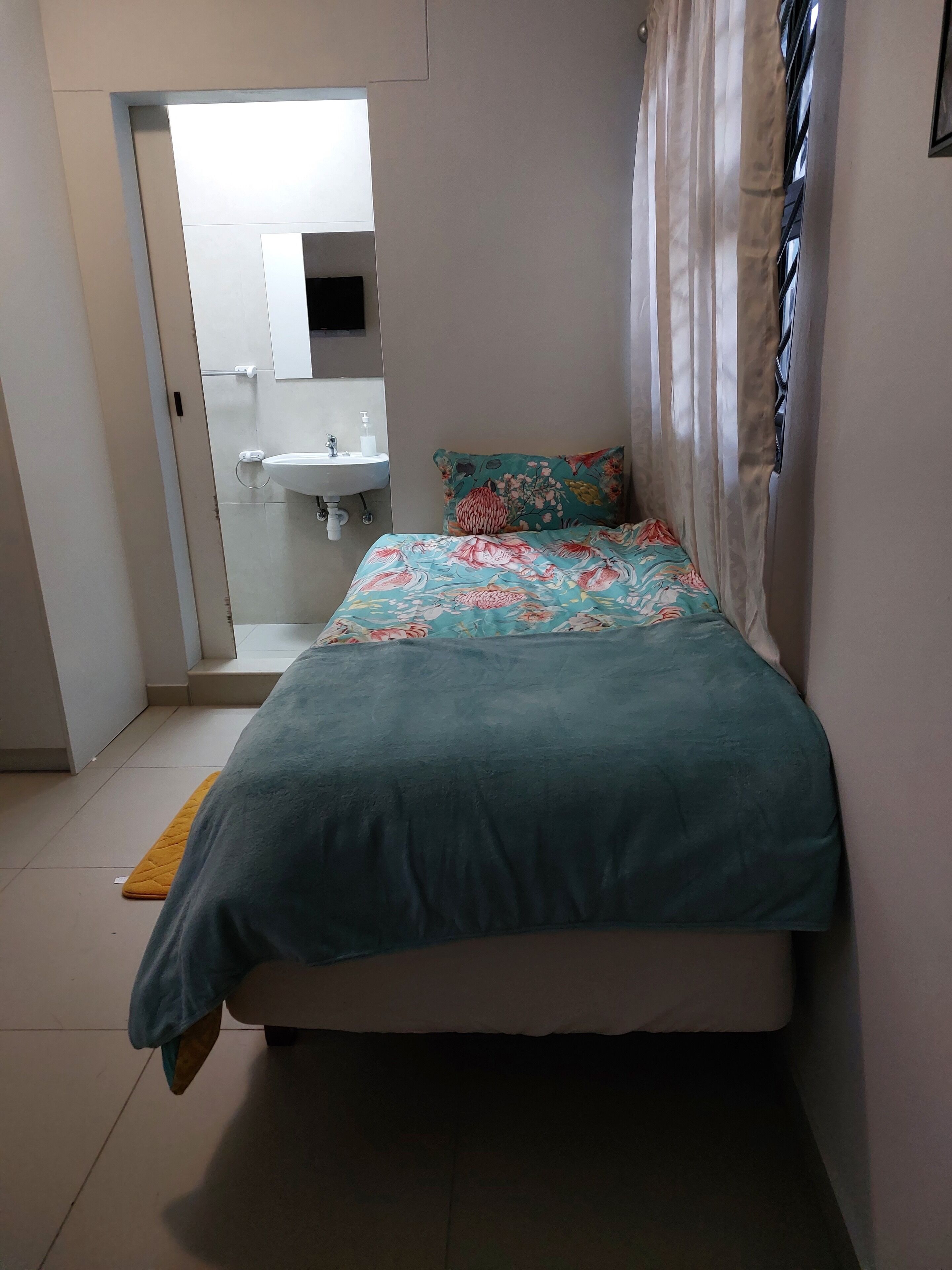 A small, neatly arranged bedroom with a bed, a bedside table, a window with curtains, and a bathroom visible through an open door.