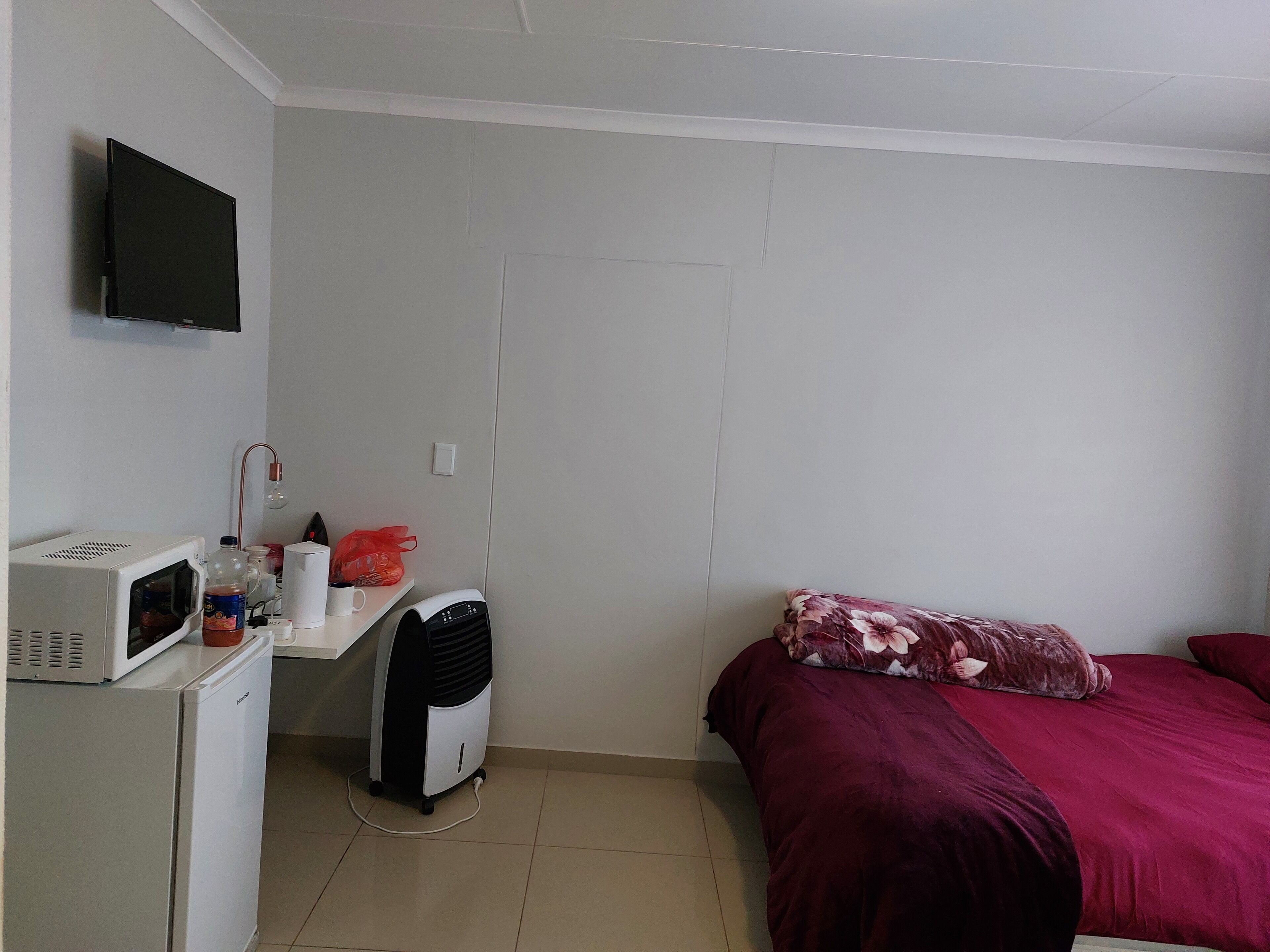 A small, compact room with a bed, a desk with a microwave, a small refrigerator, and a TV mounted on the wall.