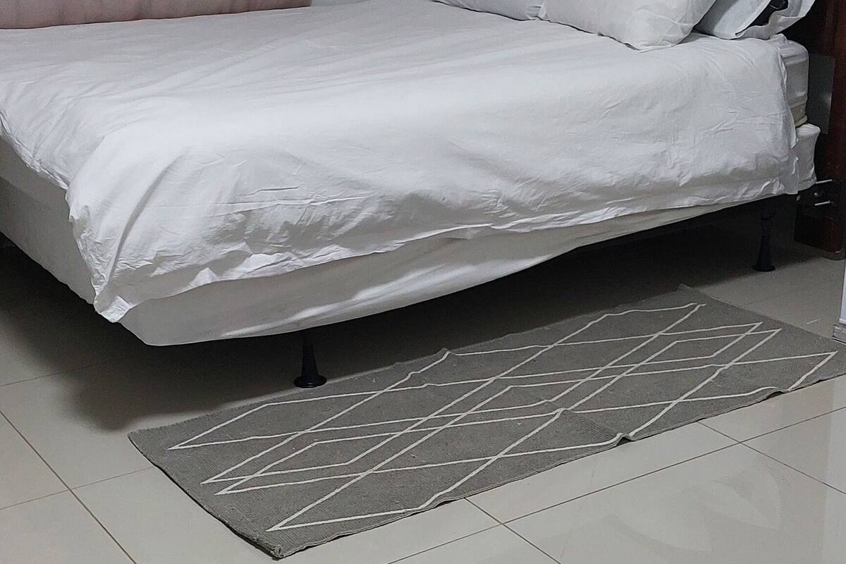 Una cama bien hecha con ropa blanca, un tapete gris estampado y un cabecero de madera.