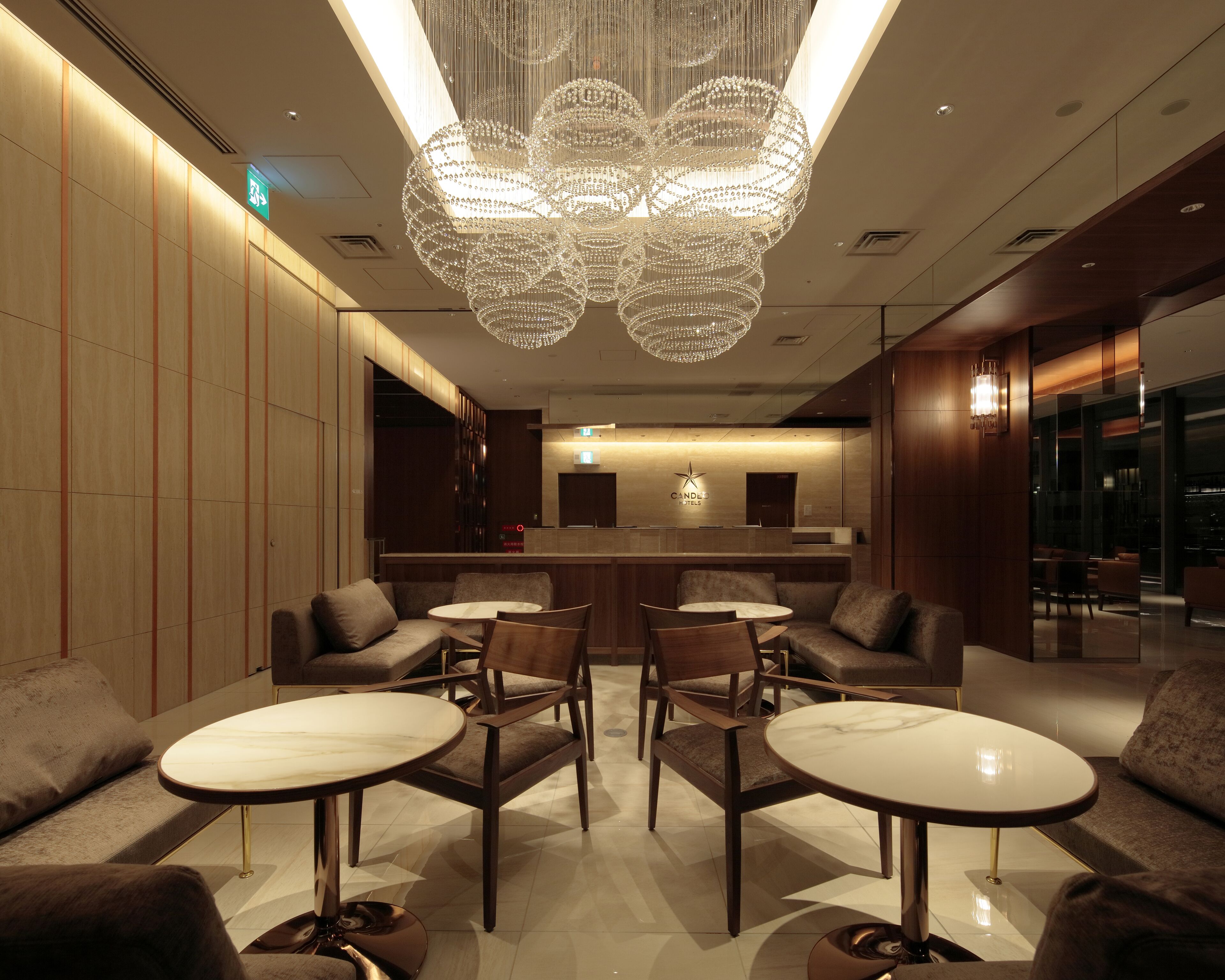 Een moderne hotellobby met een centraal receptiegedeelte, een zithoek met ronde tafels en stoelen, en een decoratieve kroonluchter.