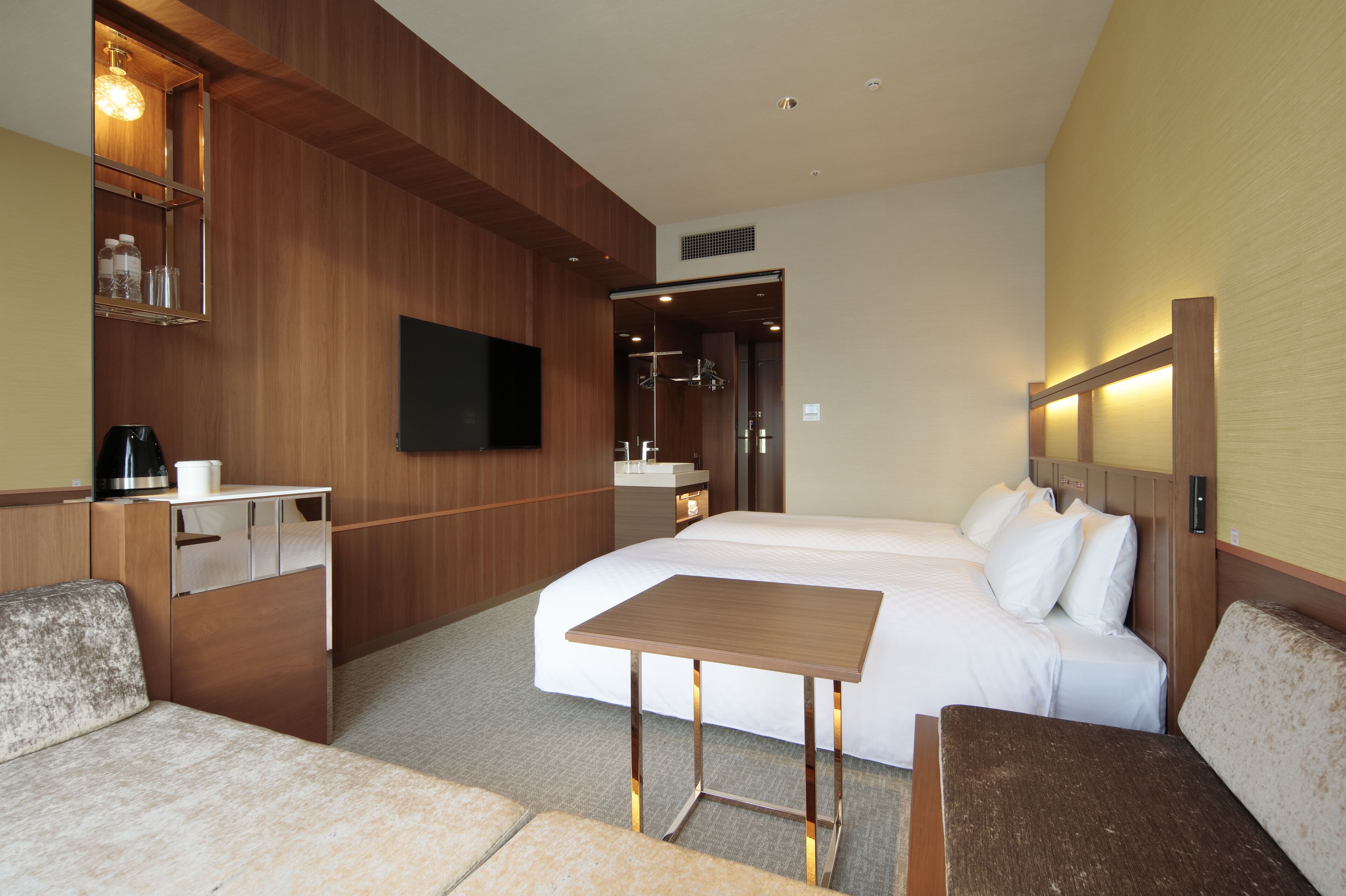 A hotel room with a bed, a small table, a sofa, a TV, and a bathroom visible through an open door.