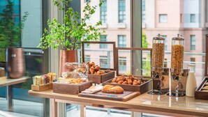 Un buffet de petit-déjeuner proposant divers plats, une plante en pot et une vue sur les bâtiments par la fenêtre.