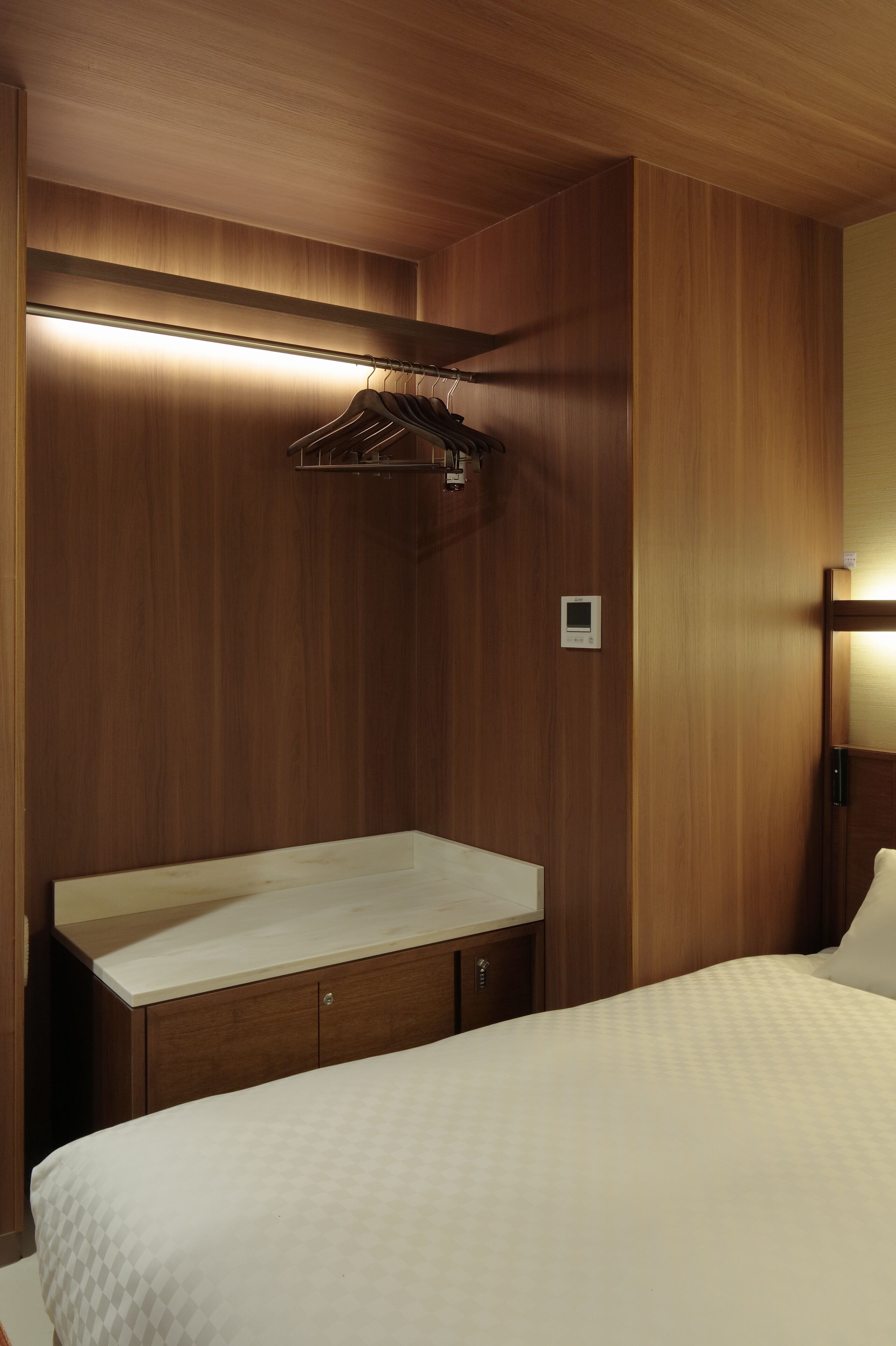 A hotel room with wooden panel walls, a bed with a white quilt, a wooden vanity with a mirror, and a hanging rack.
