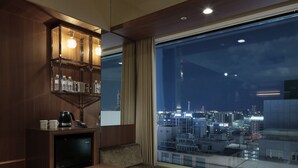 Une chambre d’hôtel moderne dotée d’une grande fenêtre donnant sur la ville, d’une table en bois, d’un canapé et d’un mini-bar intégré.
