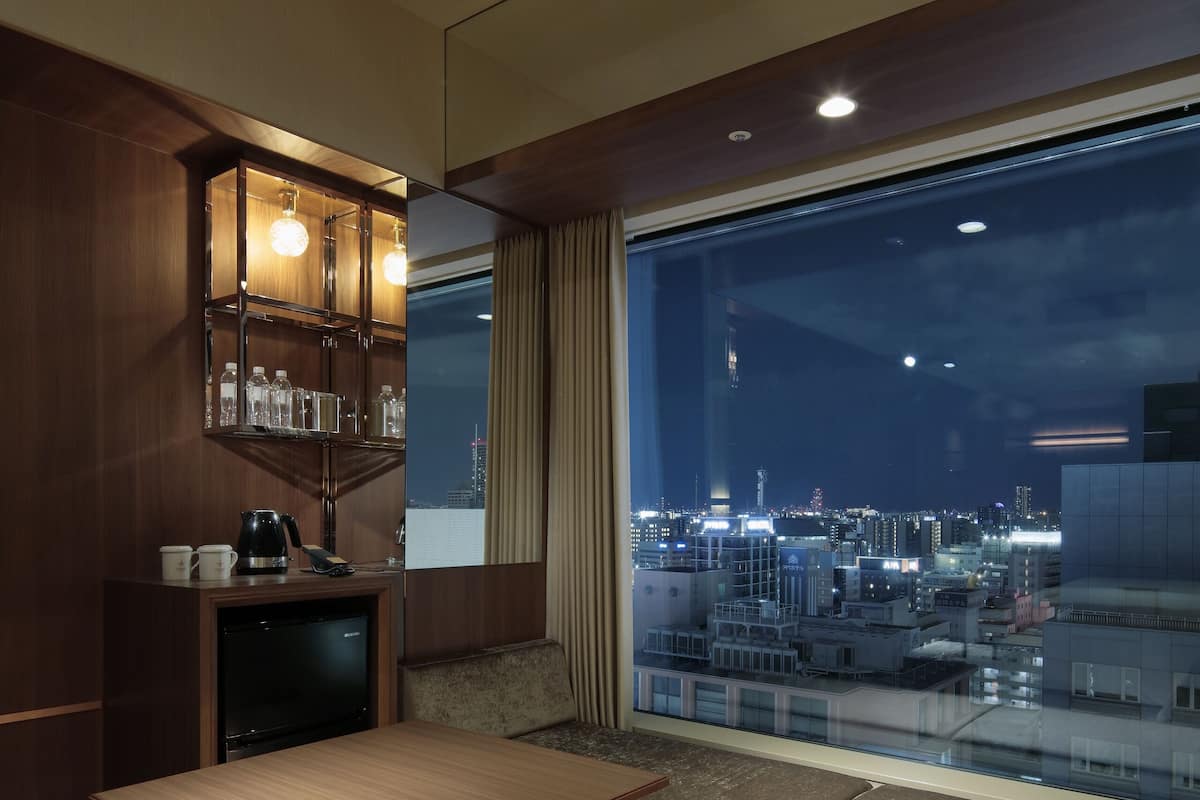 Habitación de hotel moderna con un ventanal grande que ofrece vistas a la ciudad, una mesa de madera, un sofá y un minibar empotrado.