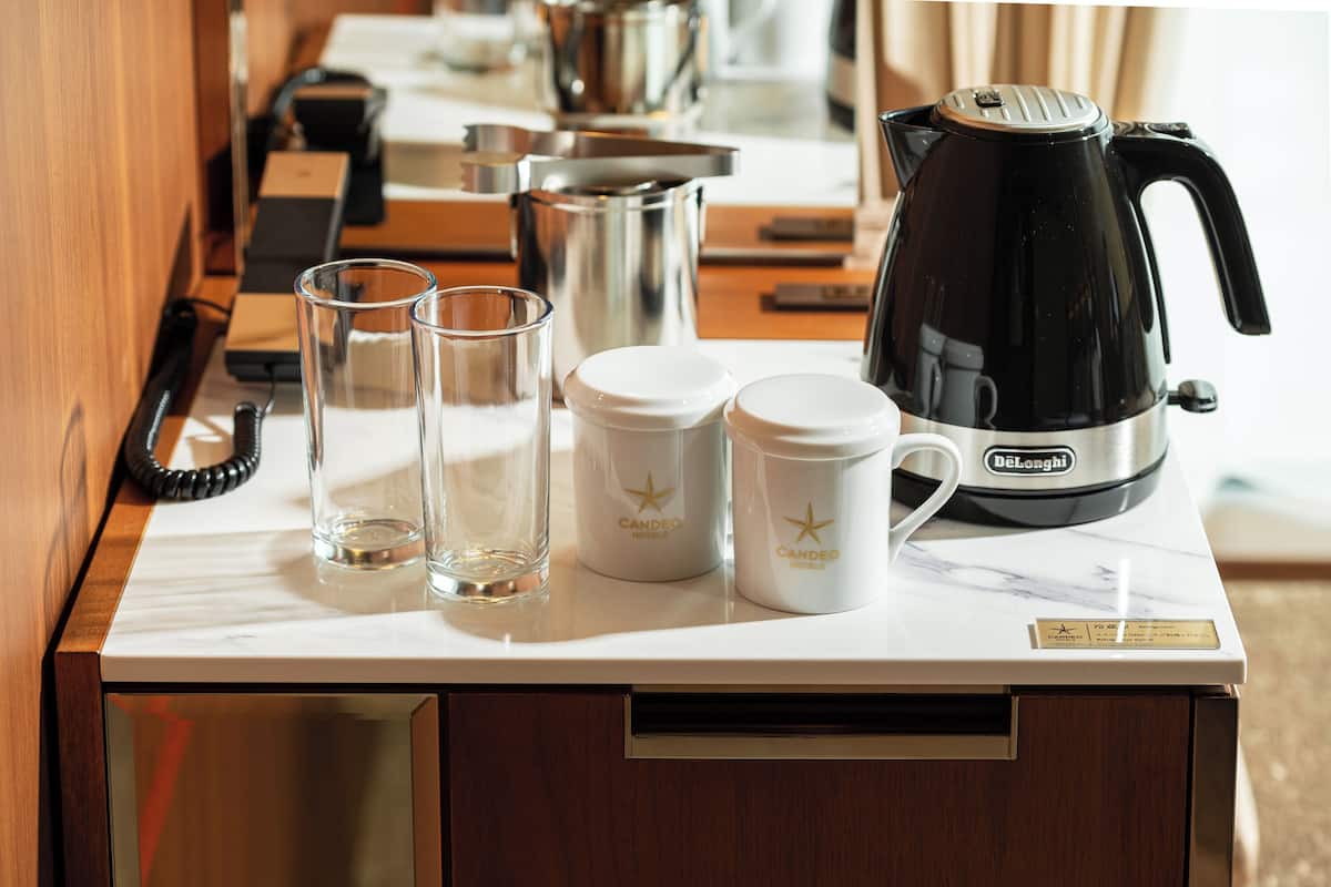 Una encimera de cocina con un hervidor eléctrico DeLonghi, dos vasos de vidrio y dos recipientes blancos con el logotipo del Hotel Candeo.