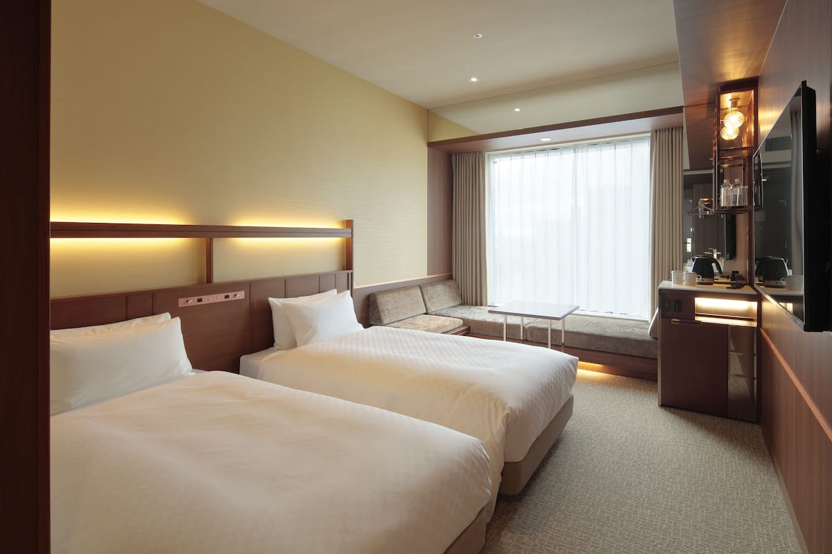 Habitación de hotel con dos camas, un pequeño espacio para sentarse, un escritorio con lámpara y un ventanal con cortinas.