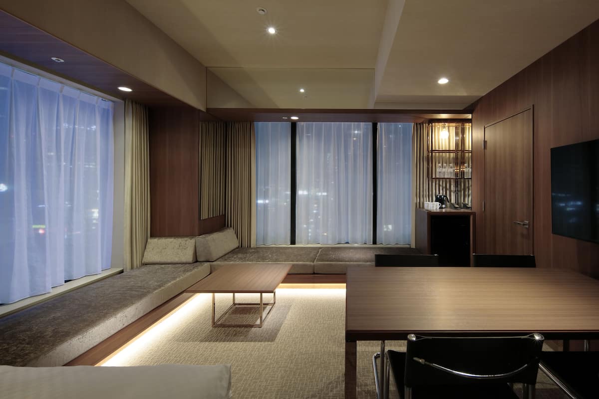 Una habitación de hotel moderna con un ventanal grande, un sofá, una mesa de comedor y un televisor.