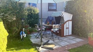 Une terrasse avec une table et des chaises, protégée par un grand parasol, entourée de verdure et agrémentée d’une petite structure surmontée d’une marquise à rayures.
