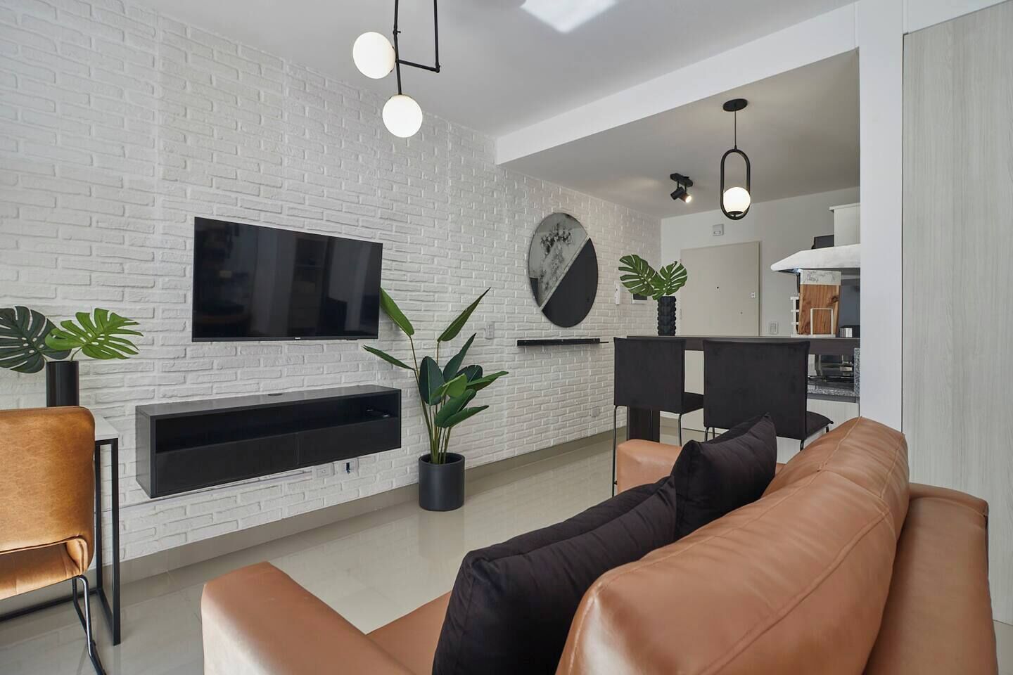 Una sala de estar moderna con una pared de ladrillo blanco, un televisor de pantalla plana, un mueble de entretenimiento negro, un sofá de cuero marrón y una zona de comedor con una mesa y sillas negras.