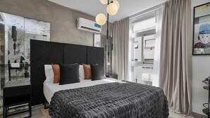 A modern bedroom with a large bed, a desk, and a chair. There is a window with curtains, an air conditioning unit, and a painting on the wall. - NoMa Work & Travel - 7A (Centro)