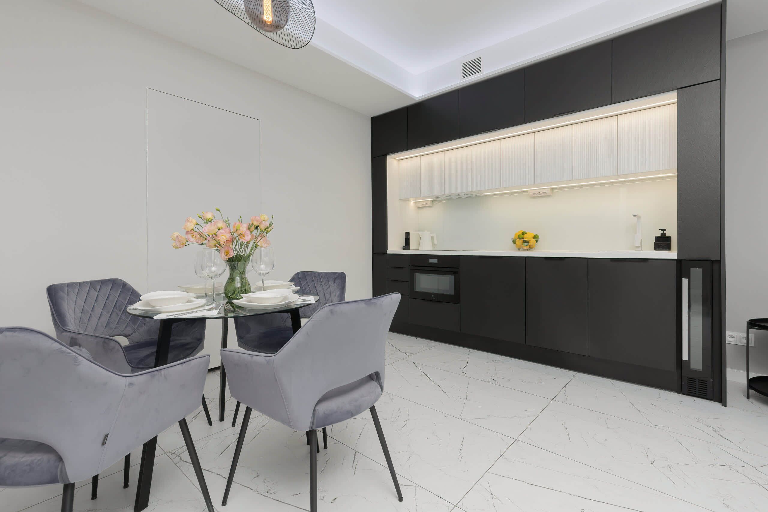 Una cucina moderna con mobili scuri, un piano di lavoro bianco e un'area pranzo con sedie grigie e un tavolo in vetro.