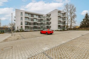 A modern apartment building with multiple balconies, a paved parking area, and a red car parked in front.
