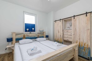 A bedroom with a wooden bed, a window with blinds, a bedside table with a lamp, and a wooden wardrobe with a curtain.