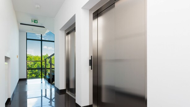 A modern elevator with a metallic finish, a glass door, and a visible emergency exit sign.