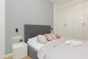 A modern bedroom with a grey headboard, a white bedside table with a plant, and a neatly made bed with rolled towels.