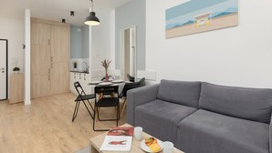 A modern living room with a grey sofa, a dining area with a table and chairs, and a kitchen in the background.