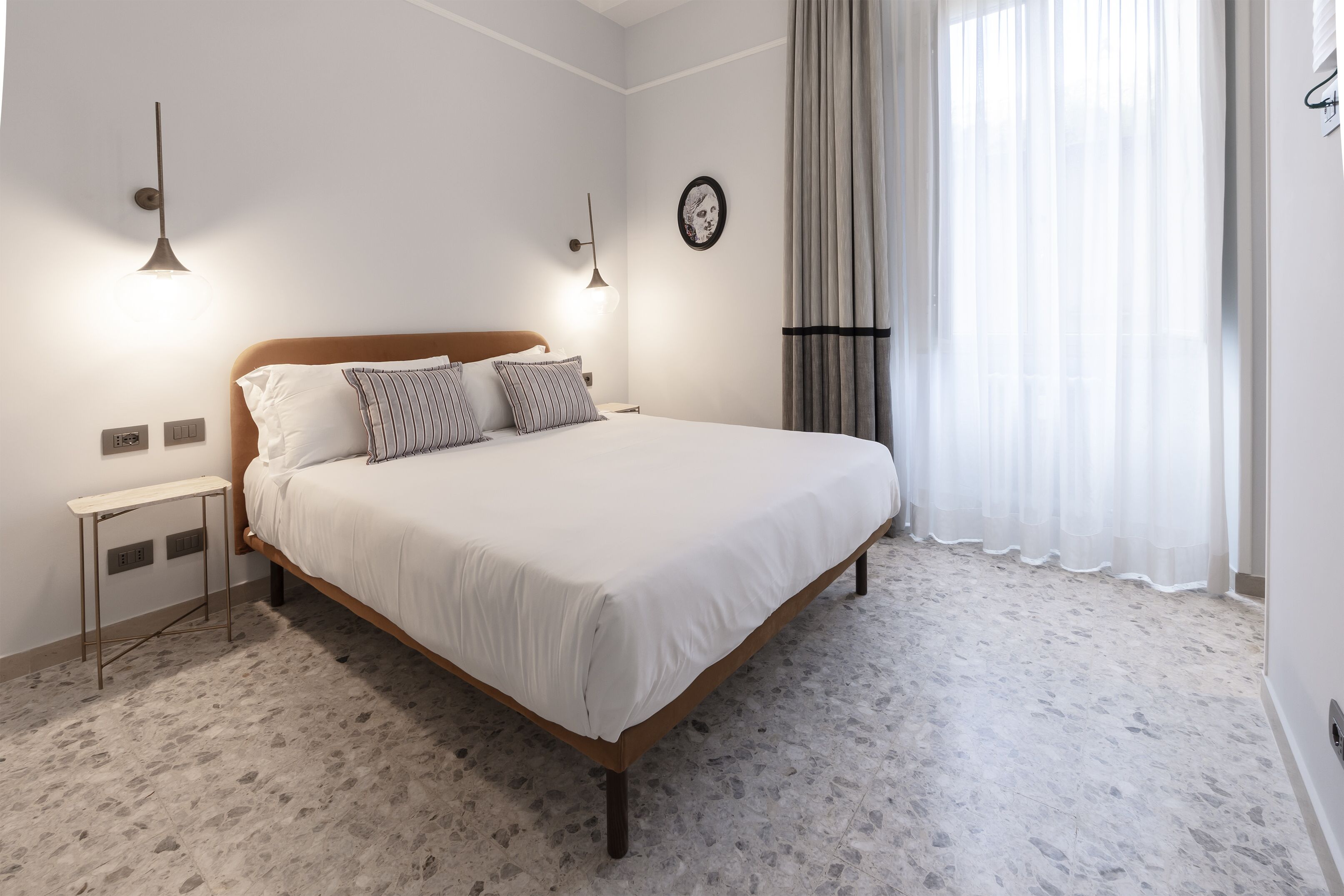 Une chambre à coucher bien rangée, avec un lit, des tables de chevet, une horloge murale et une fenêtre ornée de rideaux légers.