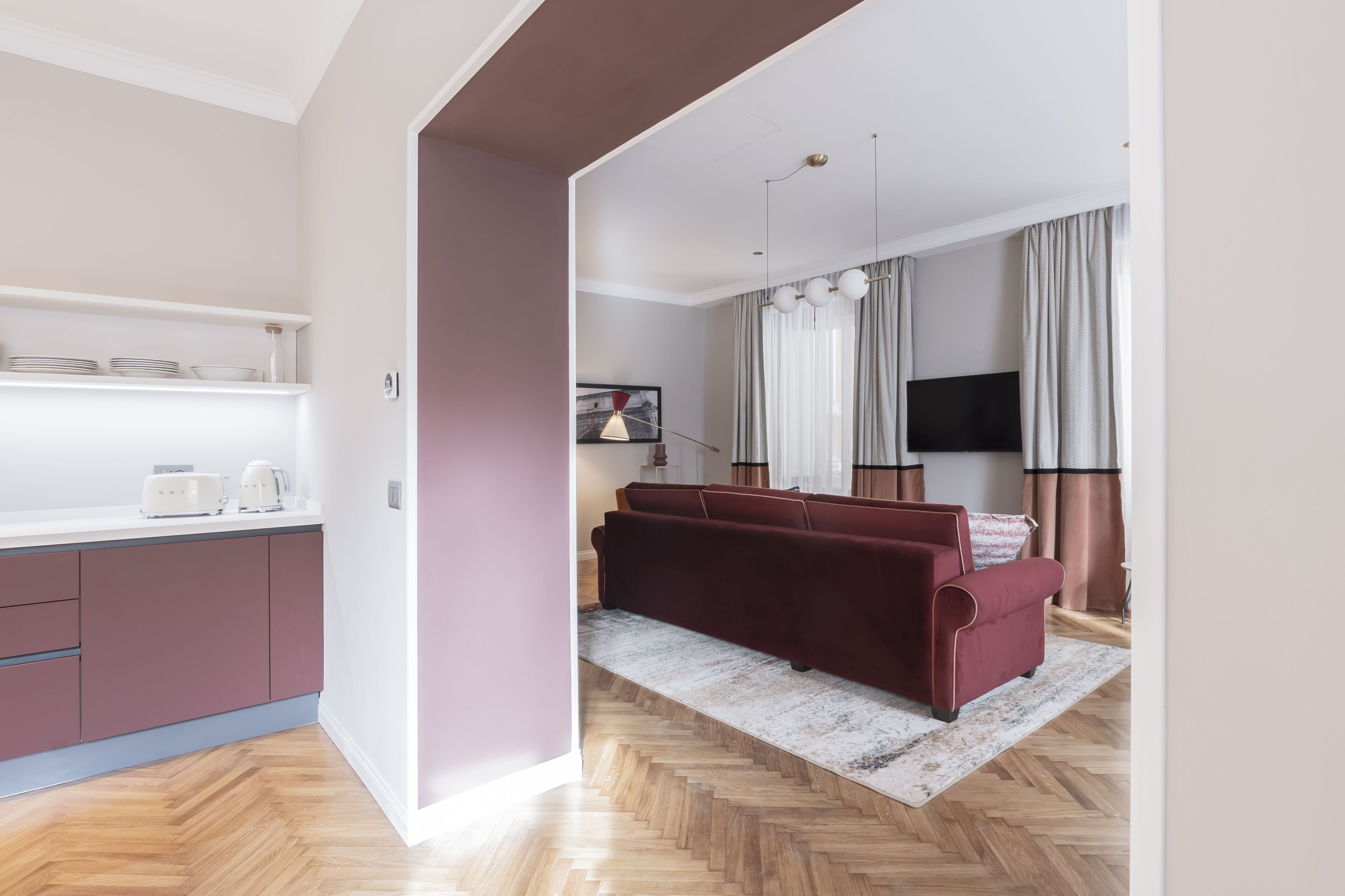A modern living room with a wooden herringbone floor, a red sofa, a flat-screen TV, and a dining area with a white countertop and purple cabinets.