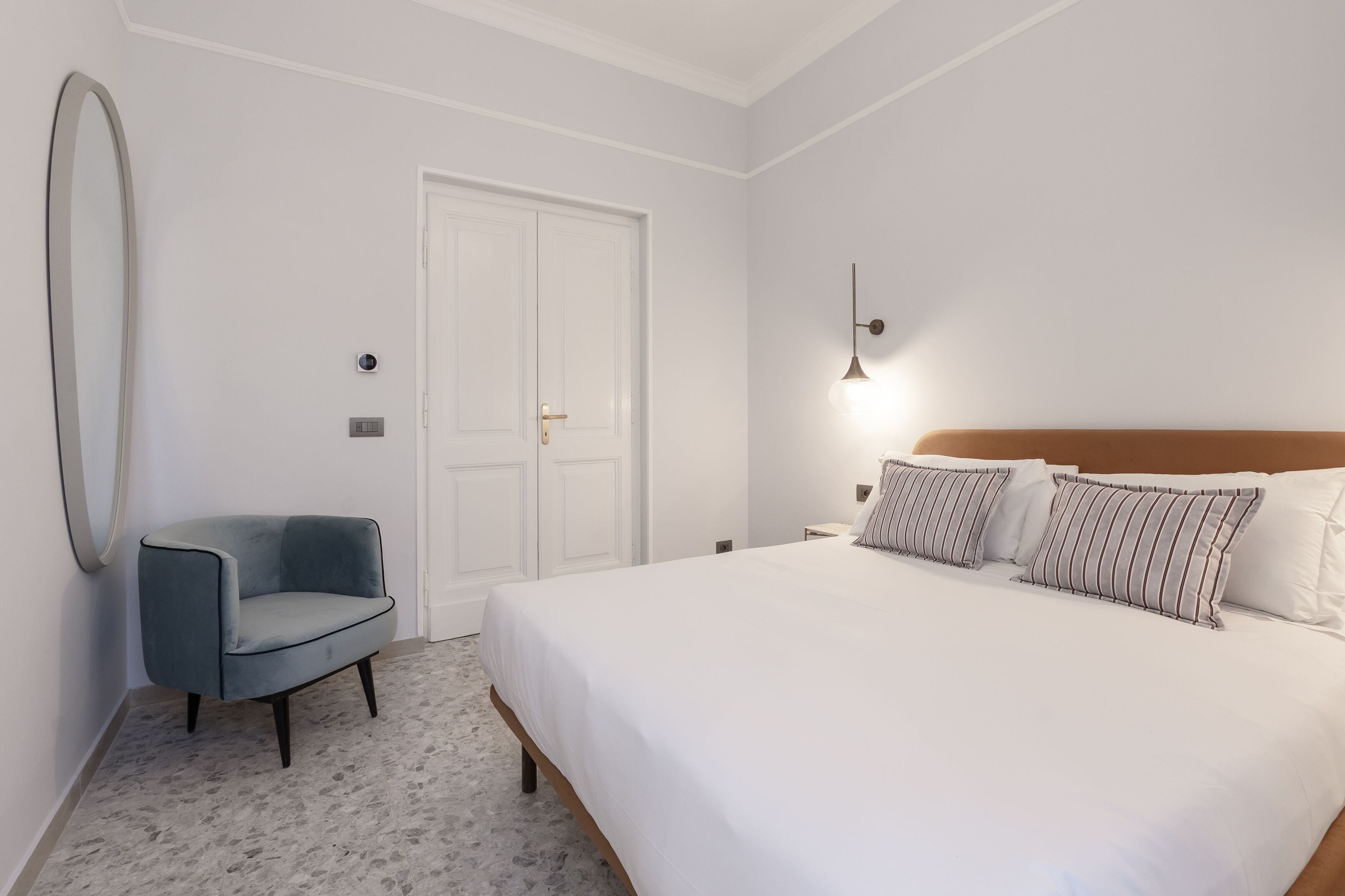 A bedroom with a bed, a chair, a door, and a wall-mounted light fixture.
