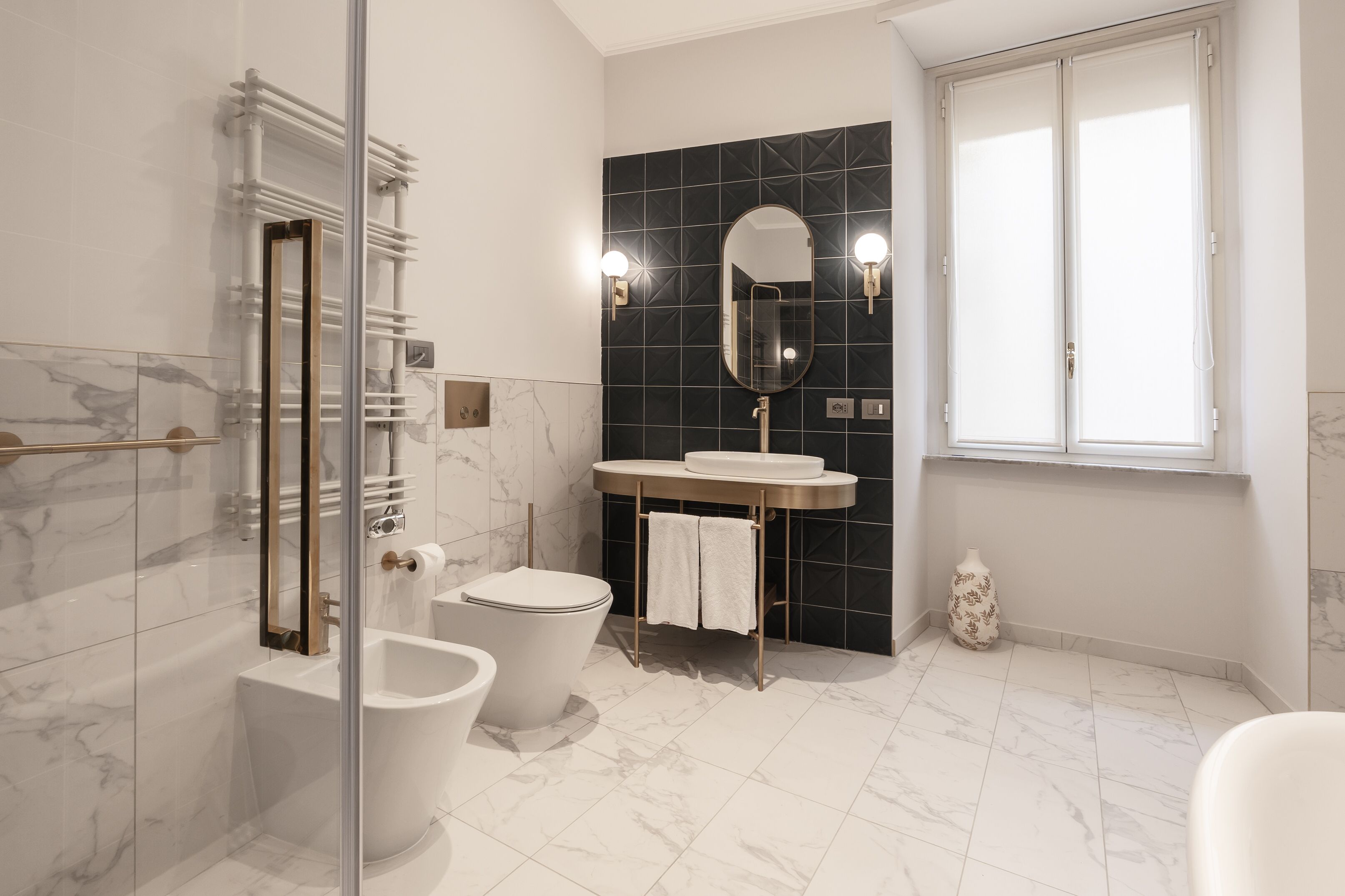 A modern bathroom with a glass shower enclosure, a toilet, a bidet, a sink with a mirror, and a window.