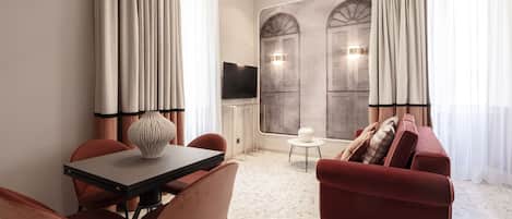 Una habitación de hotel moderna con una mesa de comedor, sillas rojas, un sofá rojo, un televisor y un ventanal con cortinas.
