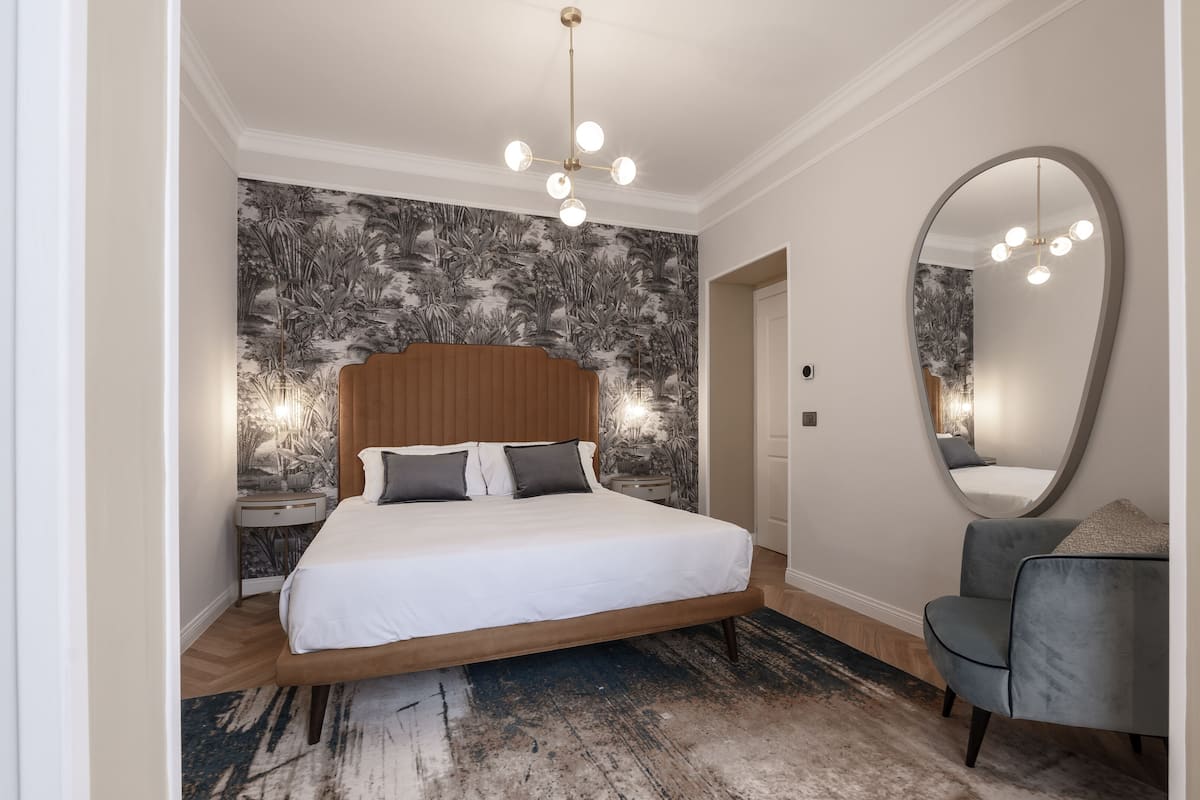Une chambre à coucher avec un grand lit, une tête de lit en bois, une table de chevet, un miroir et un fauteuil gris.