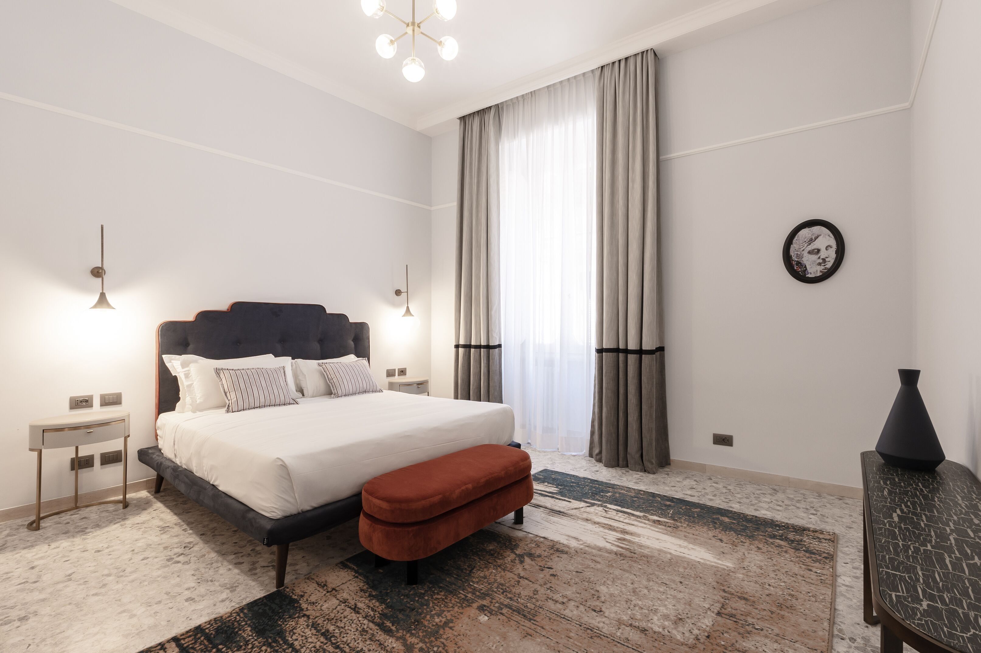 A modern hotel room with a large bed, a small bedside table, a nightstand, a bench, and a decorative vase.