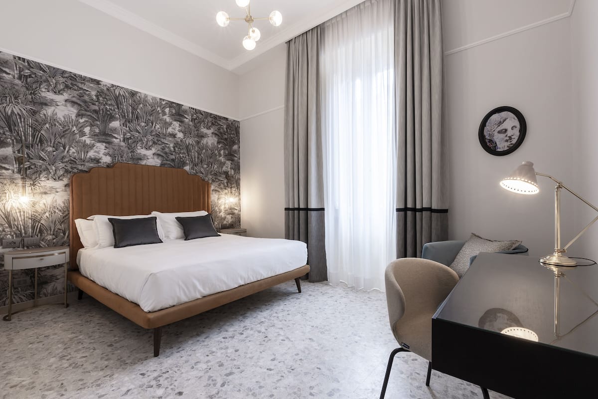 Une chambre d’hôtel moderne avec un grand lit, un bureau avec une lampe, une chaise et un mur recouvert d’un papier peint aux motifs tropicaux.