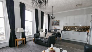 A modern living room with a black leather sofa, a dining table with a fruit bowl and a magazine, and a kitchen with white cabinets and a black backsplash. - Sunbeach (Weymouth)