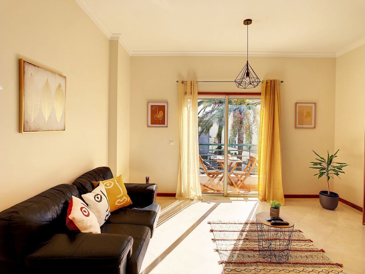 Un salon avec un canapé en cuir noir, un tapis à motifs et une vue sur un balcon aménagé avec du mobilier d’extérieur.