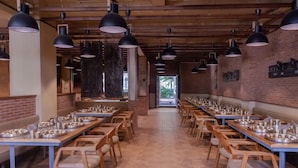 A restaurant with long communal tables and individual chairs, wooden beams on the ceiling, and pendant lighting. - Pathikashram Nilaya (Gandhinagar)