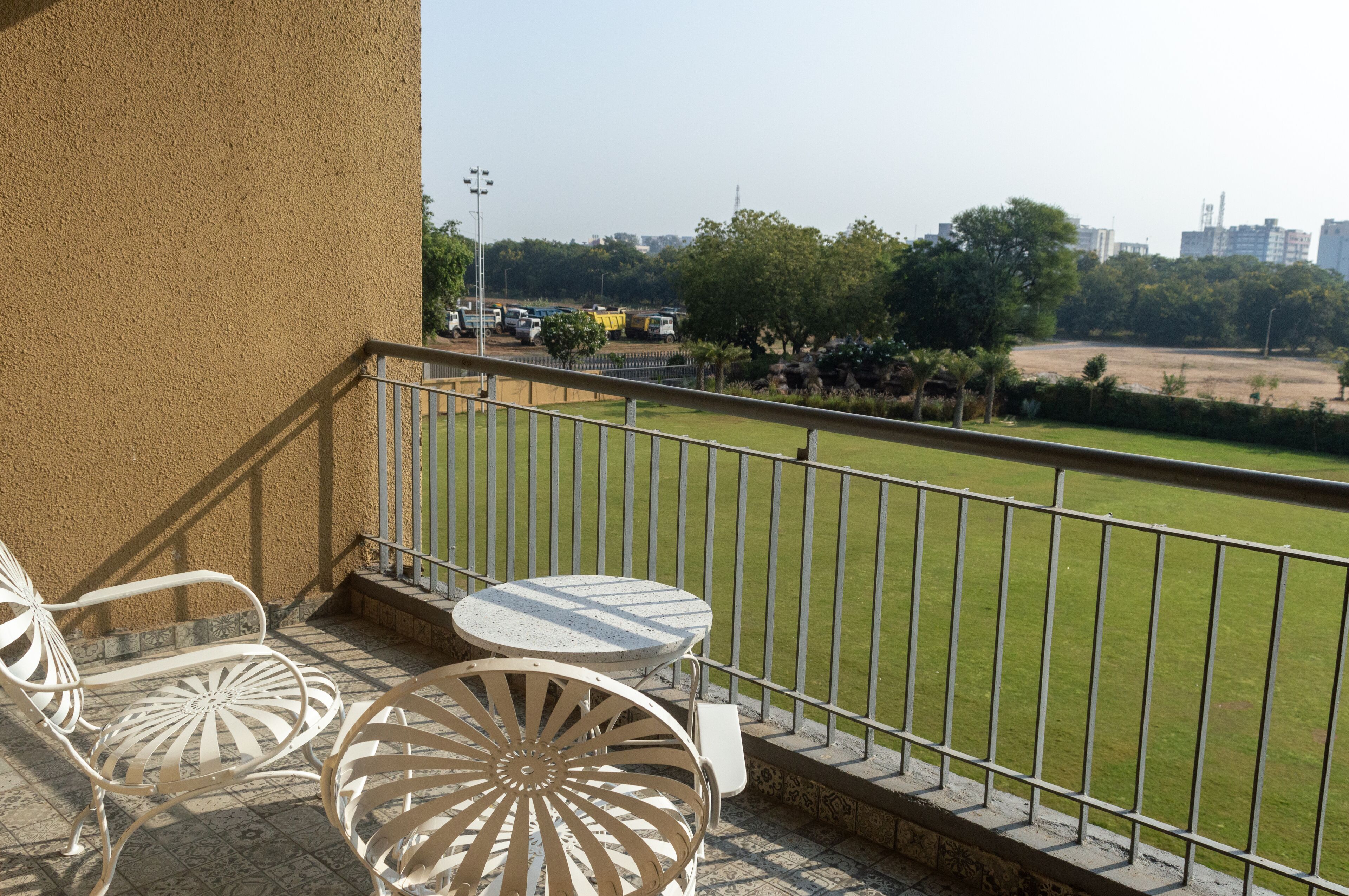 A balcony with two white metal chairs and a small round table, overlooking a grassy field and a parking area with several vehicles.