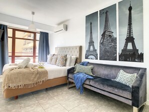 Quarto de hotel com uma cama, um sofá, uma janela com cortinas e três posters da Torre Eiffel na parede.